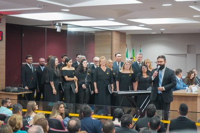 Homens e mulheres de preto no palco e o maestro ao lado do teclado, falando ao microfone, de fre...