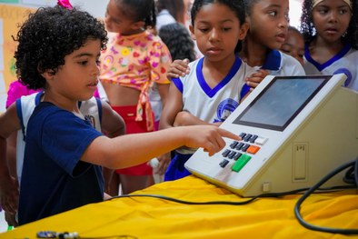 A imagem mostra uma criança sorridente, usando óculos e uma camisa branca com detalhes em azul, ...