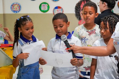 A imagem mostra uma criança sorridente, usando óculos e uma camisa branca com detalhes em azul, ...