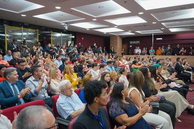 A imagem mostra o palco do plenário do TRE-RN e parte da plateia, com todos de pé, em posição de...