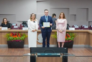 A imagem mostra o palco do plenário do TRE-RN e parte da plateia, com todos de pé, em posição de...