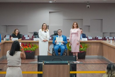 A imagem mostra o palco do plenário do TRE-RN e parte da plateia, com todos de pé, em posição de...