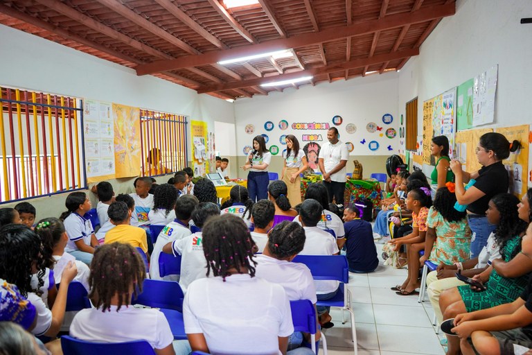 A imagem mostra um refeitório de uma escola, com os alunos sentados de costas, sentados em carte...